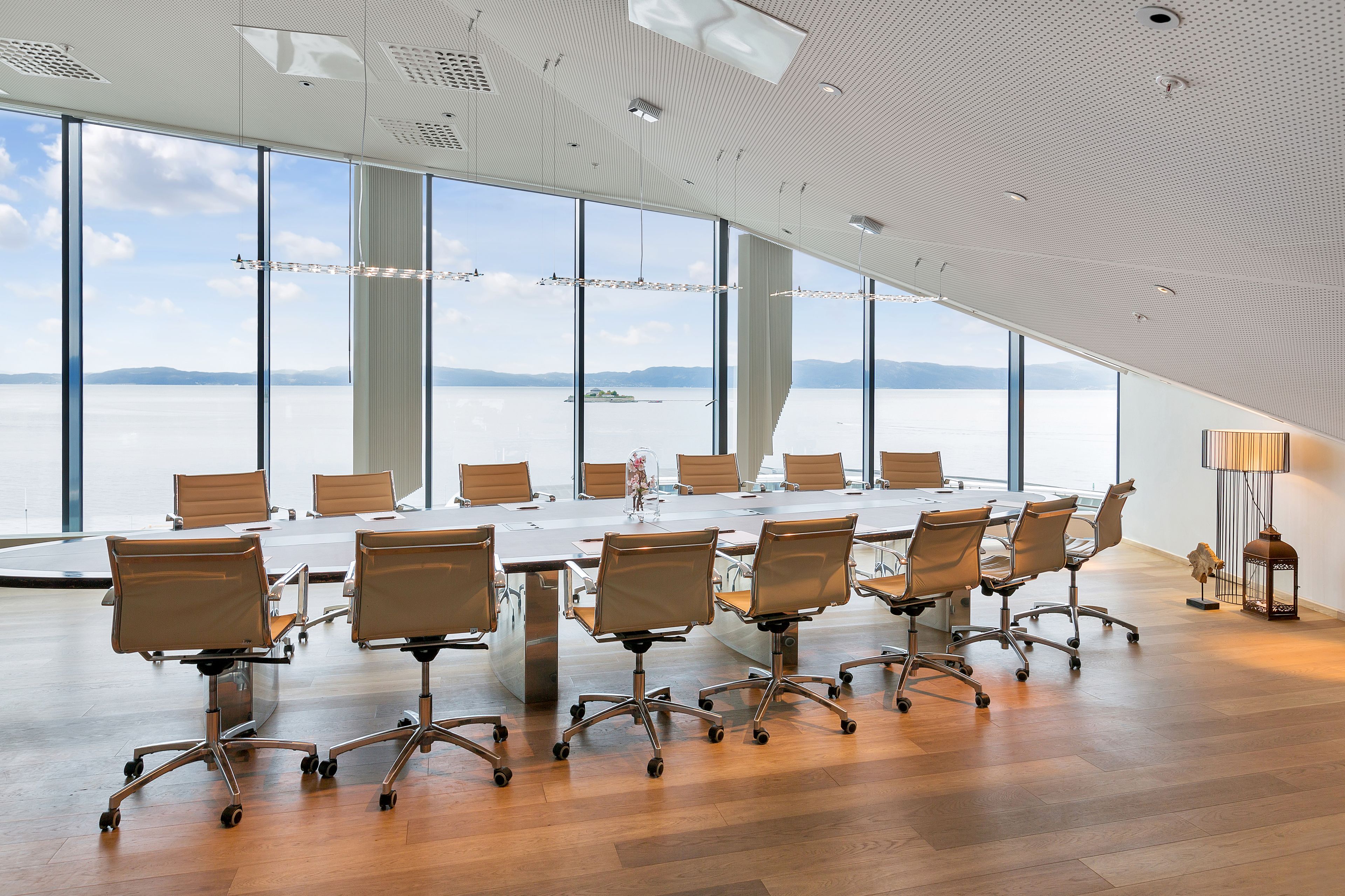 Un salón de conferencias con una gran mesa y sillas, ventanas amplias con vistas a un cuerpo de agua y montañas, y un diseño de techo moderno.