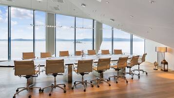 Un salón de conferencias con una gran mesa y sillas, ventanas amplias con vistas a un cuerpo de agua y montañas, y un diseño de techo moderno.