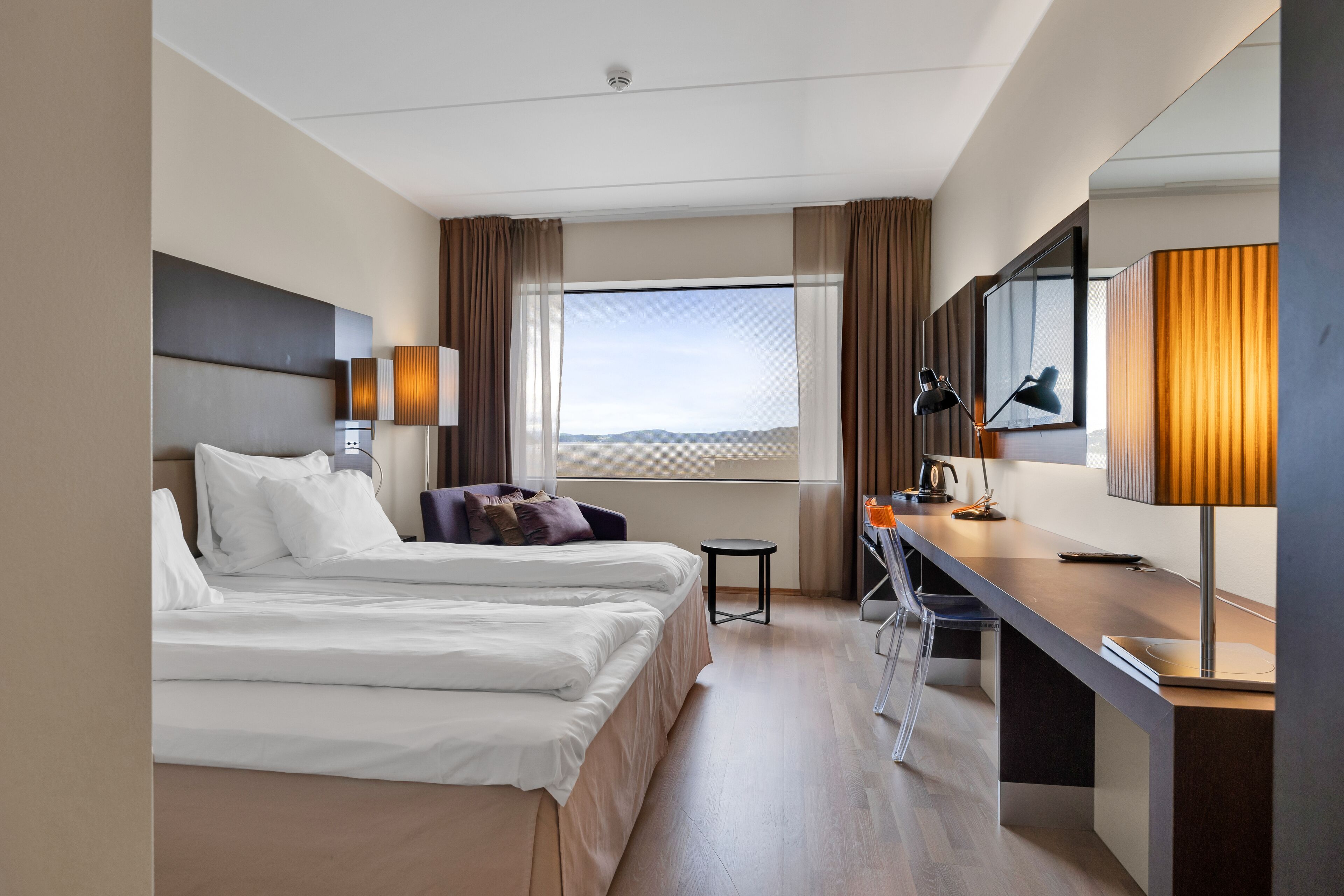 A hotel room with two beds, a desk, a chair, a small table, a mirror, and a window with a view.