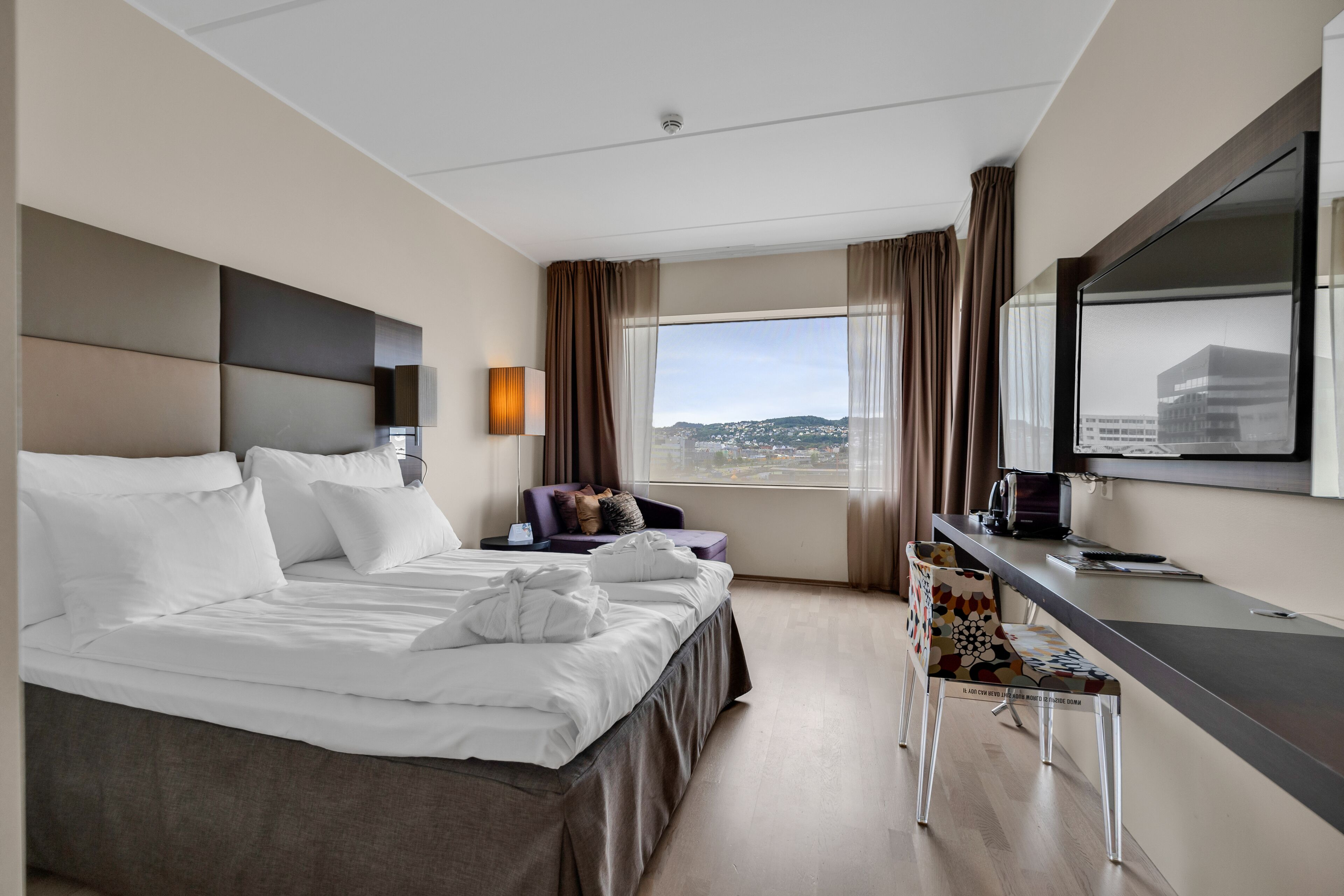 Une chambre d’hôtel moderne avec un grand lit, un bureau et une chaise. Une fenêtre offre une vue sur la ville.