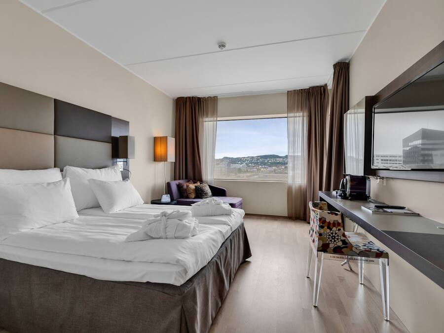 Habitación de hotel moderna con una cama grande, un escritorio y una silla. Se ve una ventana con vistas a la ciudad.