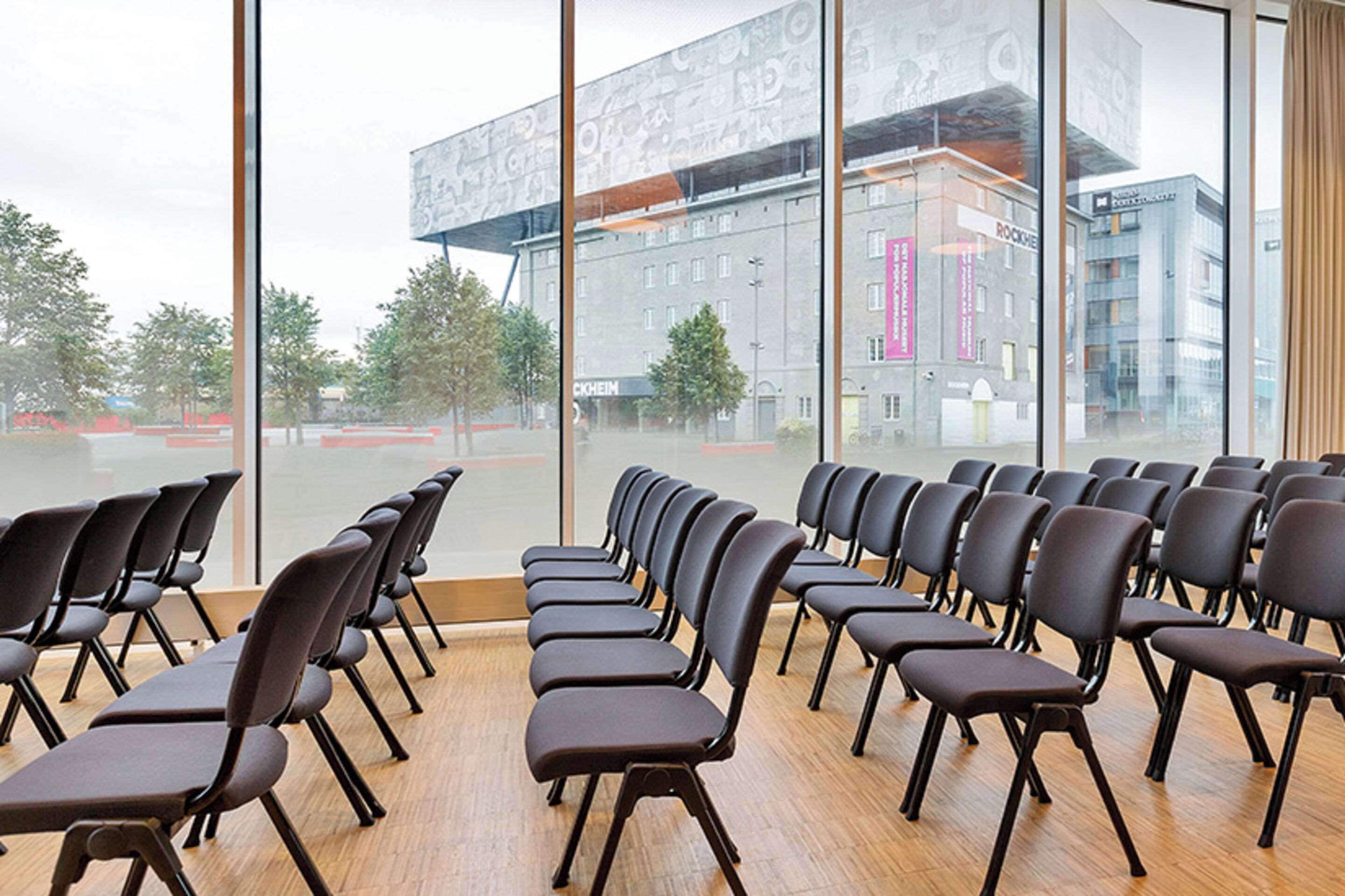 Une salle de conférence avec des rangées de chaises disposées face à une grande fenêtre donnant sur un panorama urbain.