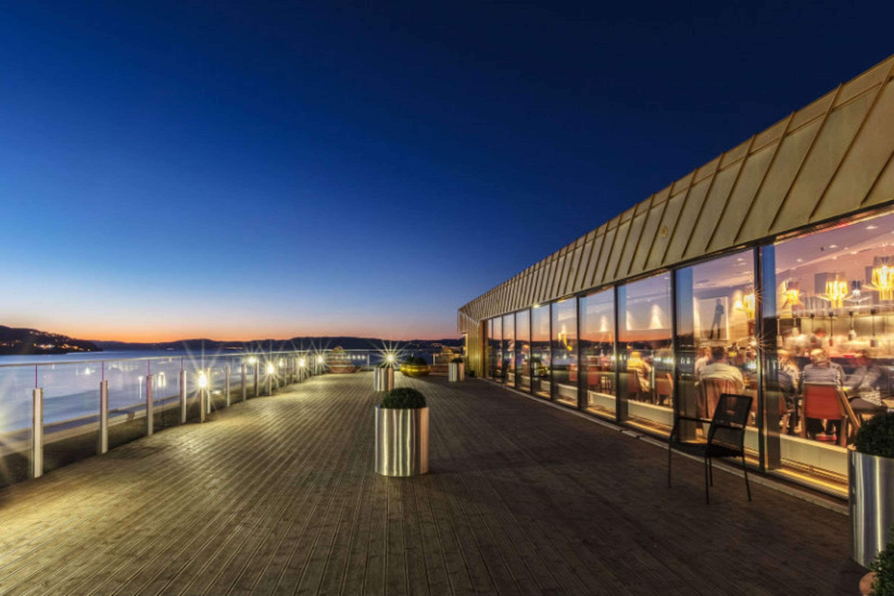 Un restaurant situé au bord de l’eau, doté d’une terrasse en bois, de grandes fenêtres et offrant une vue sur l’eau.