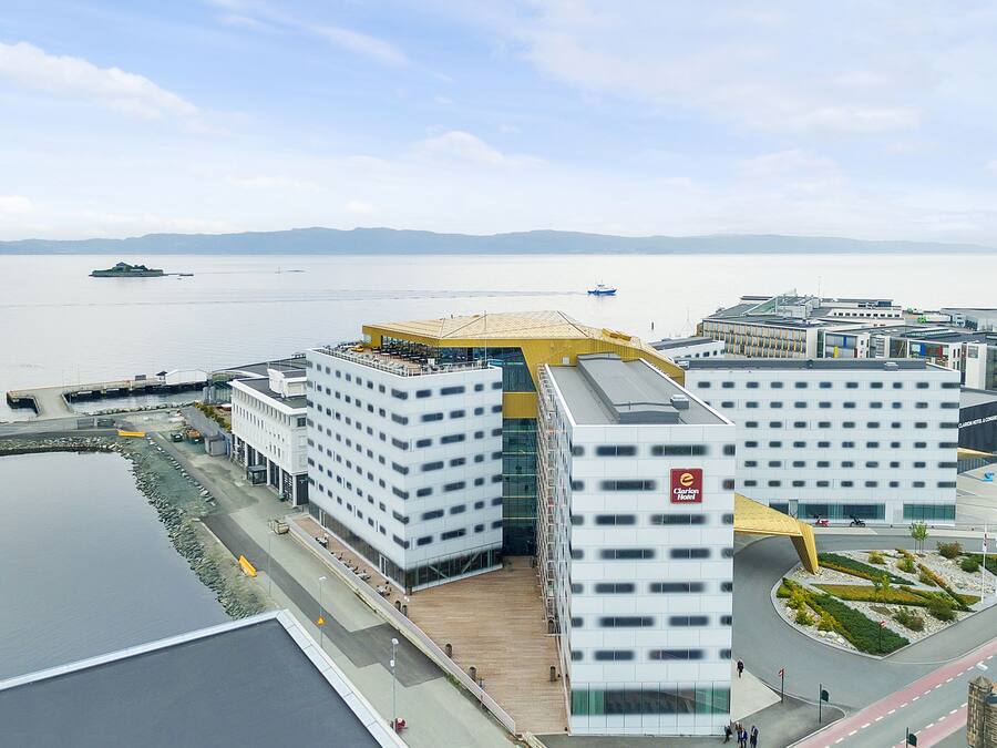 A modern hotel with a prominent yellow roof, situated by a waterfront with a view of the sea and distant mountains.