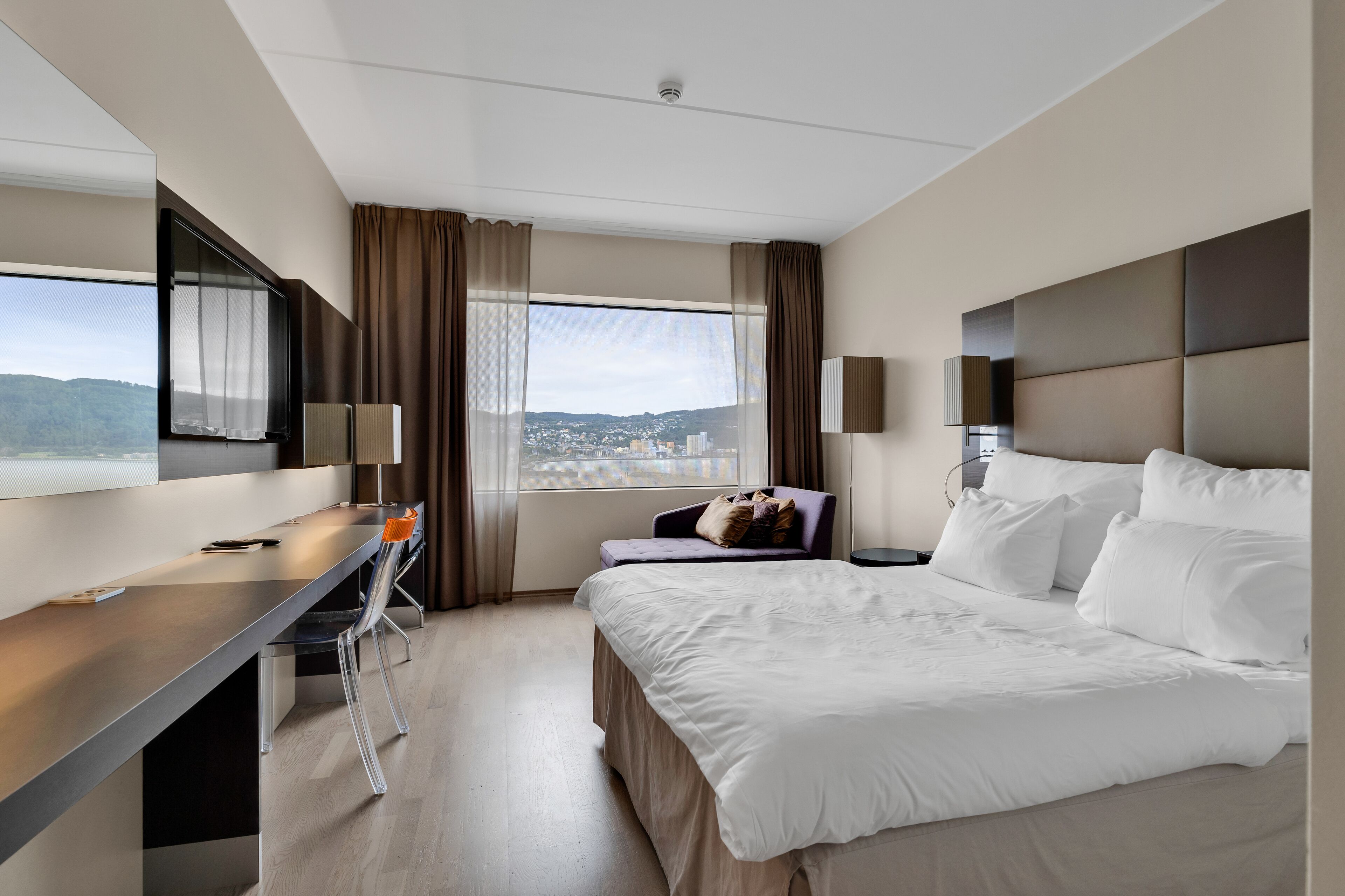 A modern hotel room with a large bed, a desk with a chair, a TV, and a view of a cityscape.