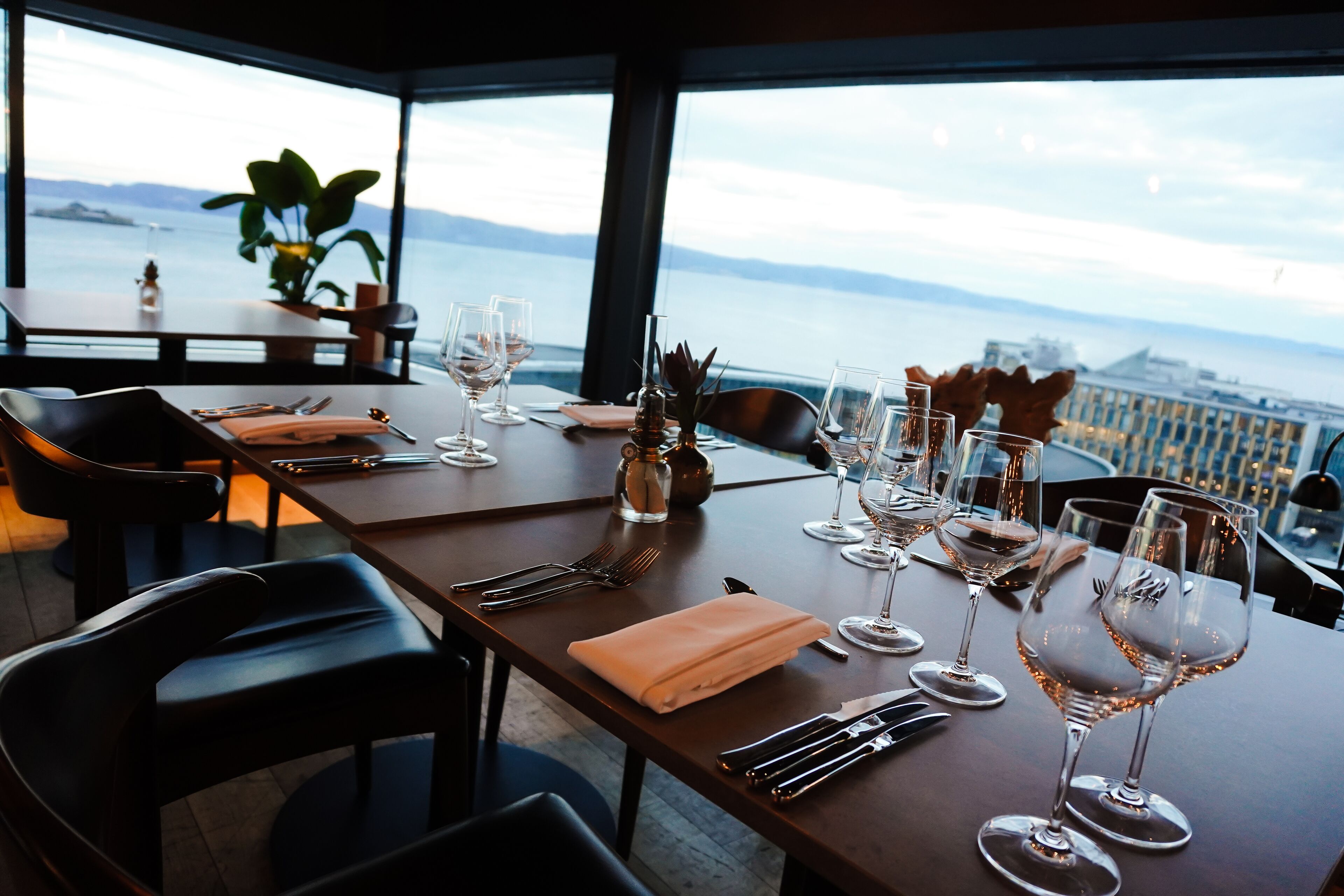 Mesa de comedor servida con copas de vino, cubiertos y una servilleta doblada, con vista panorámica al agua y las montañas.