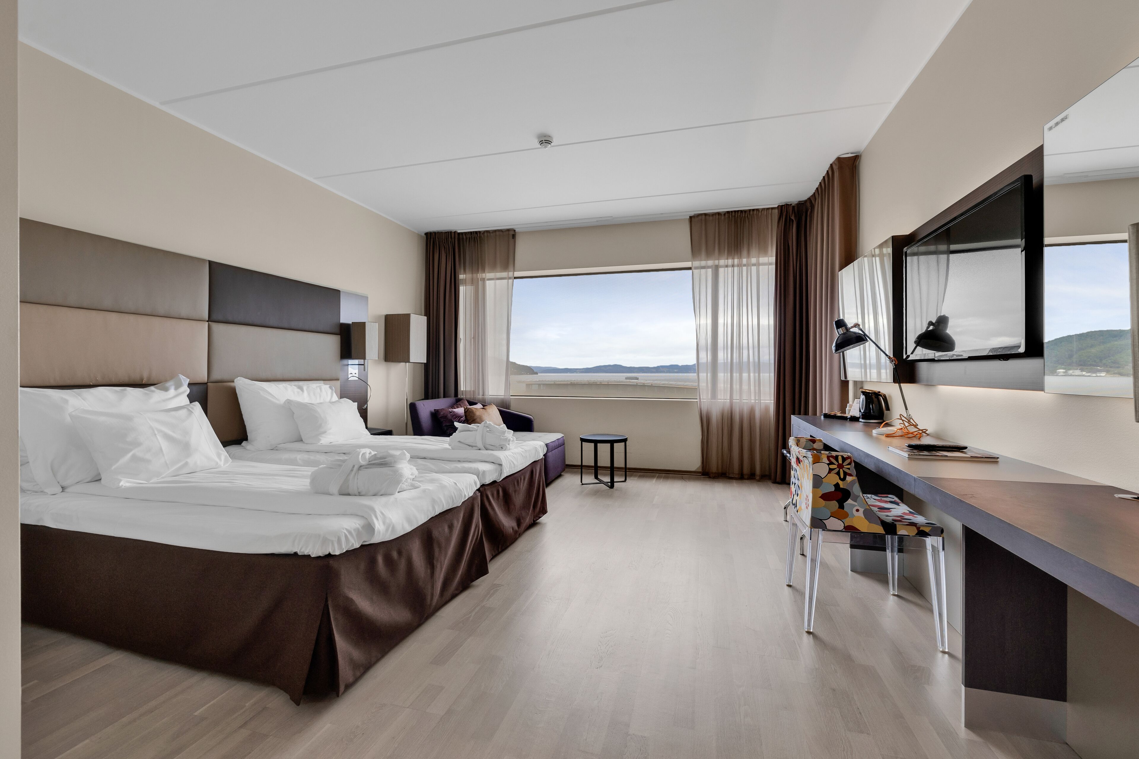 Une chambre d’hôtel avec un grand lit, un bureau, une chaise, une petite table et une fenêtre donnant sur un beau paysage.