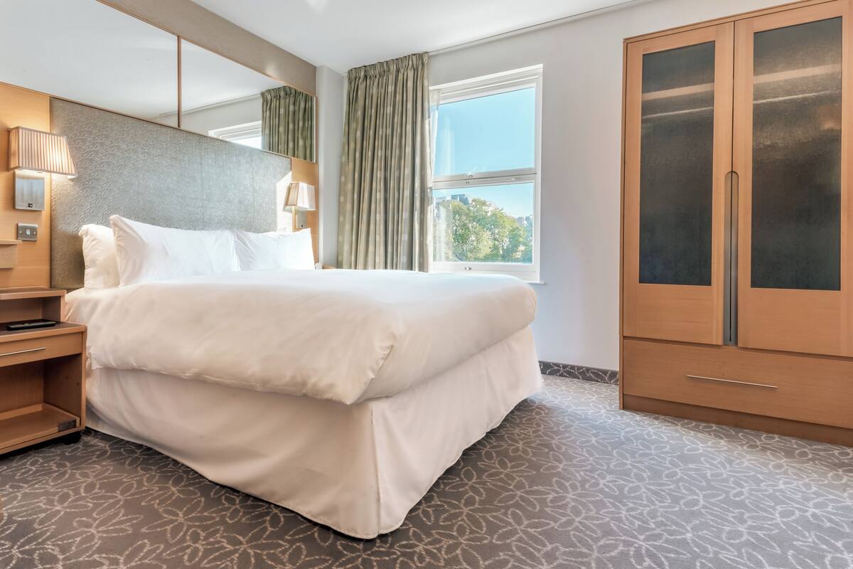 Ein Hotelzimmer mit einem großen Bett, einem Kleiderschrank, einem Nachttisch mit Lampe und einem fenster mit Vorhängen.