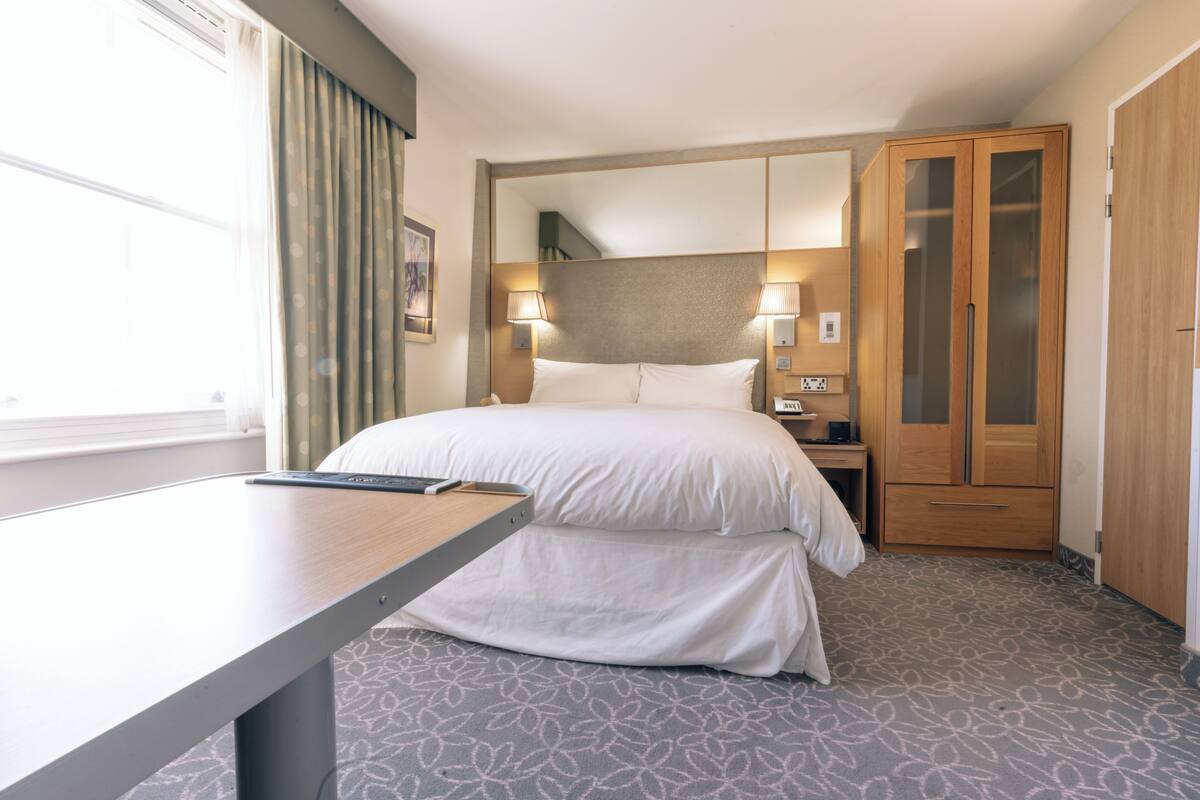 Ein Hotelzimmer mit Bett, Nachttisch, Lampe, Kleiderschrank und einem Fenster mit Vorhängen.