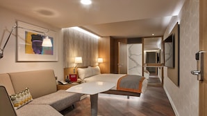 A modern hotel room with a large bed, a sofa, a dining table, and a TV mounted on the wall.