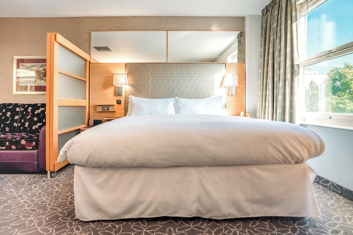 Ein Hotelzimmer mit einem großen Bett, einem Nachttisch, einer Lampe, einem Fenster mit Vorhängen und einer gemusterten Couch.