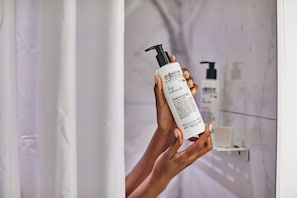 Une personne tenant un flacon d’après-shampoing Bigelow dans une salle de bain dont le mur est en marbre, avec un distributeur de savon en arrière-plan.