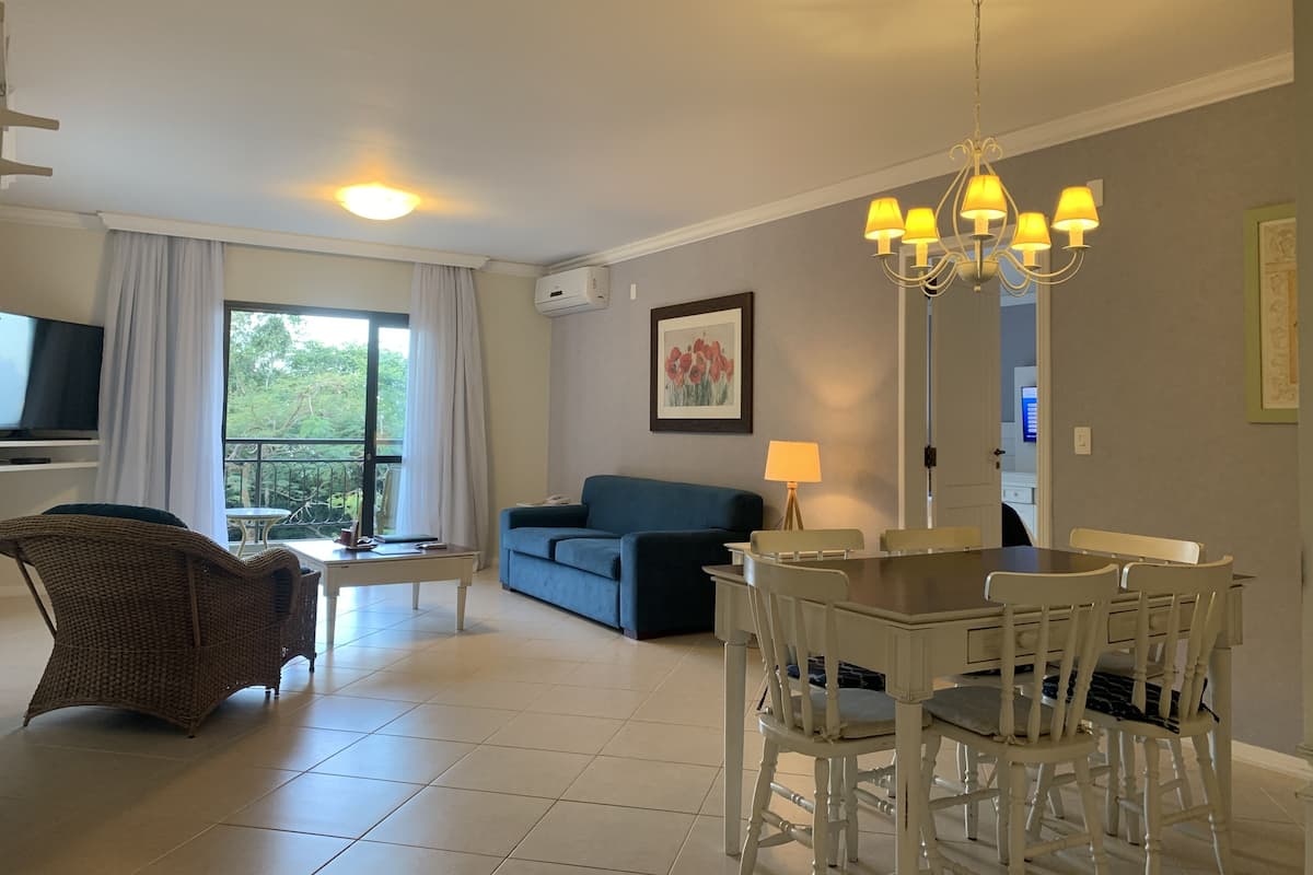 Ein Wohnzimmer mit Sofa, Esstisch, Stühlen, Fernseher und einem Balkon mit Vorhängen.