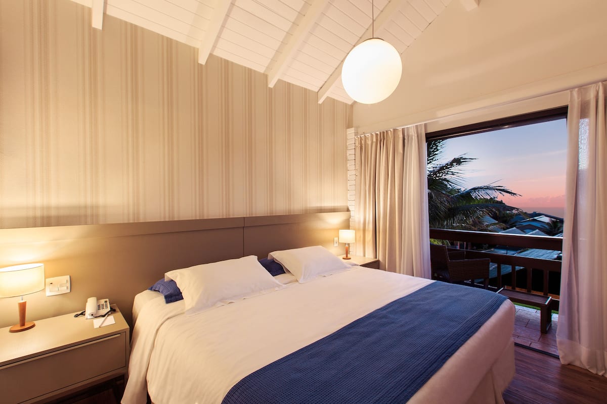 Ein Hotelzimmer mit einem großen Bett, einem Nachttisch, einer Lampe und Blick auf den Strand bei Sonnenuntergang.