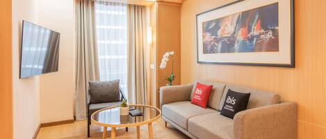 A modern hotel room with a sofa, a round coffee table, a flat-screen TV, and a framed abstract painting.