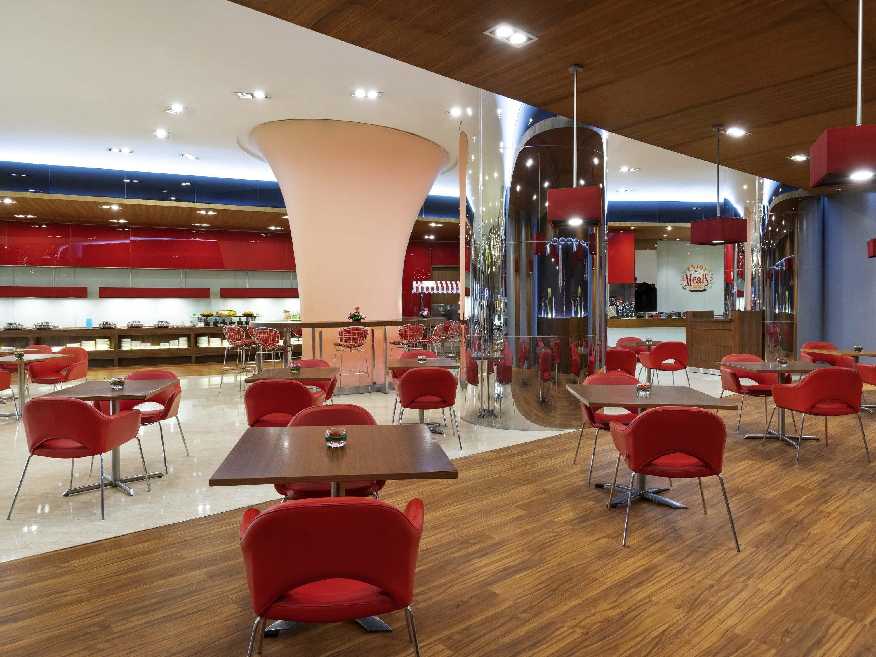 A modern restaurant with red chairs and tables, a central light fixture, and a counter with a menu.