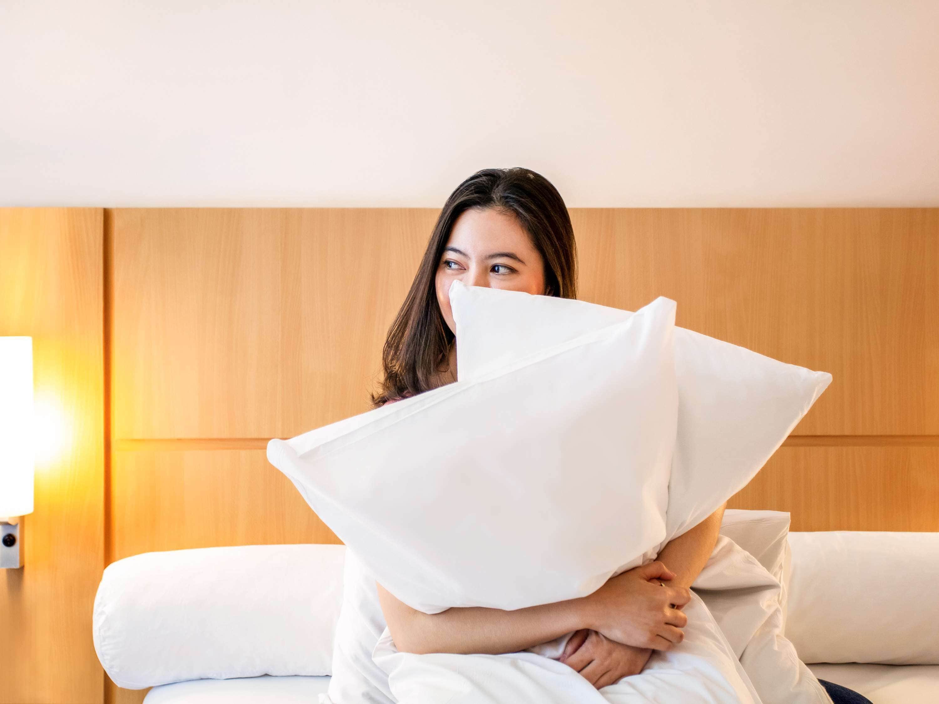 Una persona sentada sobre una cama con ropa de cama blanca, sosteniendo un almohadón blanco.