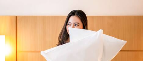 Una persona sentada sobre una cama con ropa de cama blanca, sosteniendo un almohadón blanco.