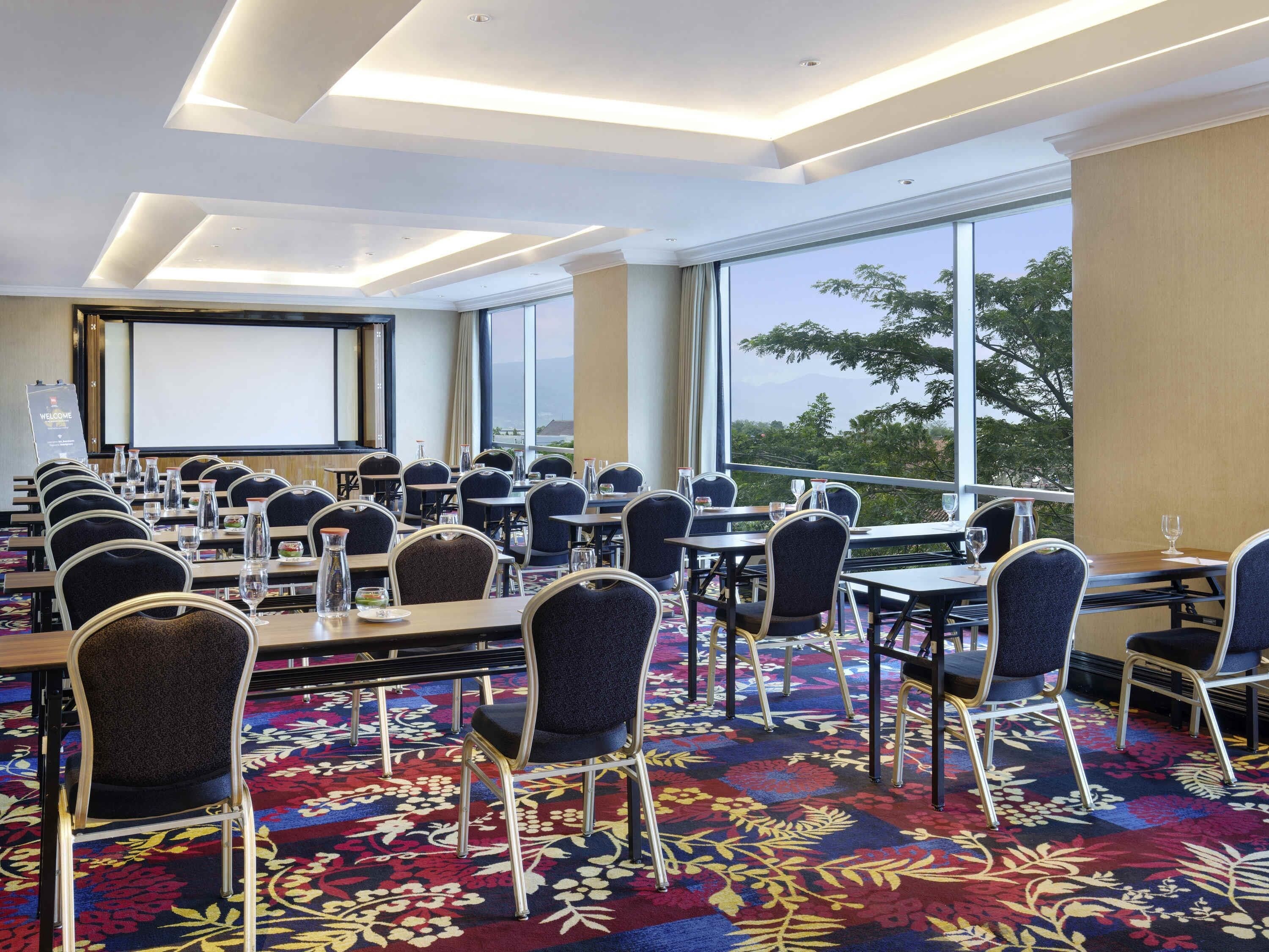 Une salle de conférence équipée de tables et de chaises, d’un écran de projection et de grandes fenêtres offrant une vue dégagée.