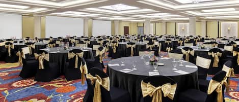 A large banquet hall with round tables, black chairs with gold sashes, and a colorful carpet.
