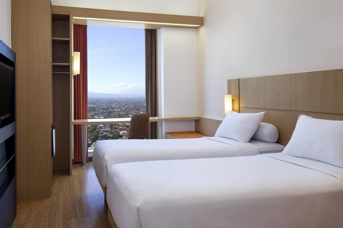 Ein Hotelzimmer mit zwei Betten, einem Fernseher, einem Kleiderschrank und einem großen Fenster mit Blick auf die Stadt.