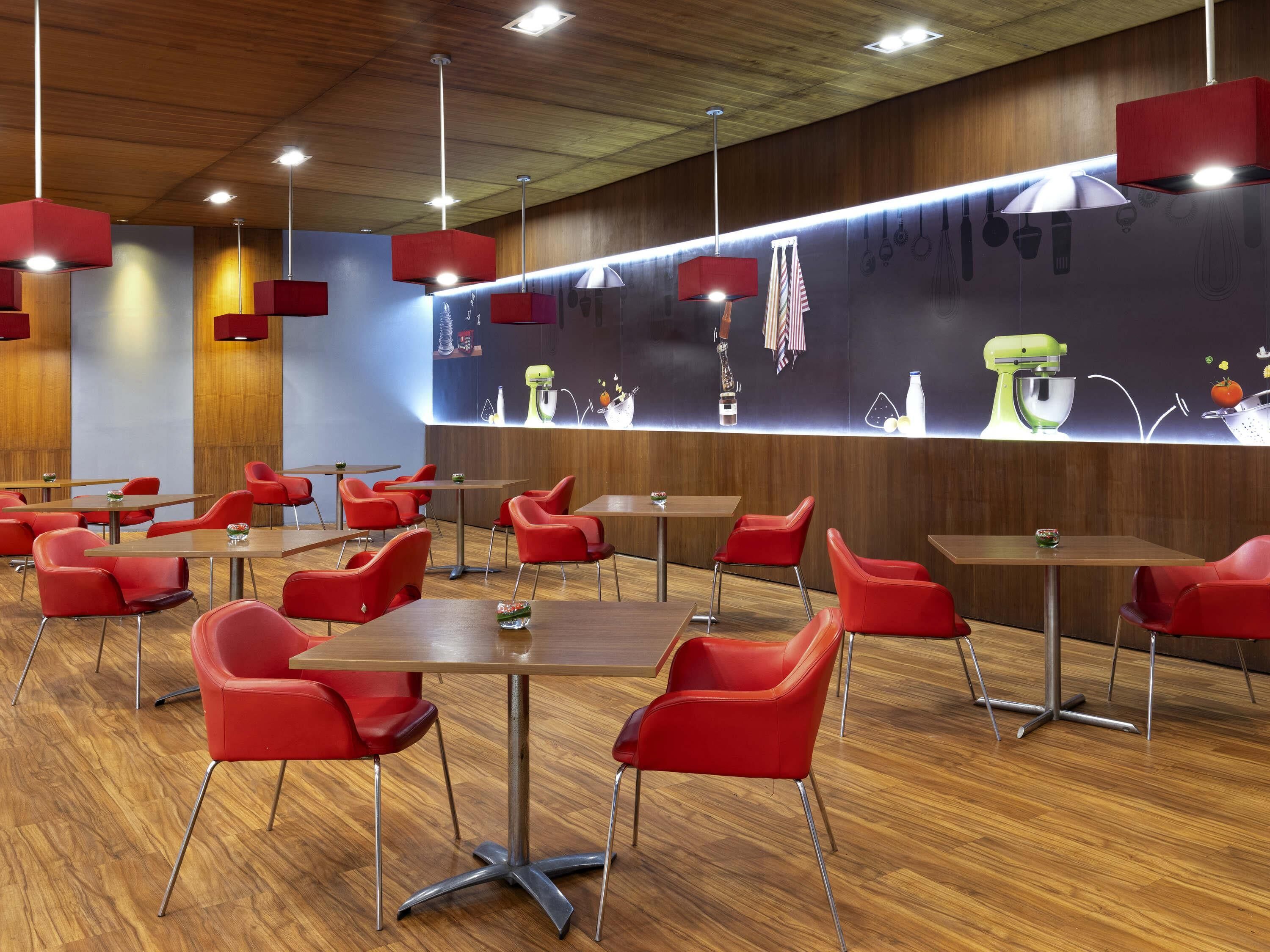 Un comedor moderno con sillas rojas y mesas de madera, con una pared temática de cocina que incluye utensilios colgantes y un electrodoméstico verde.
