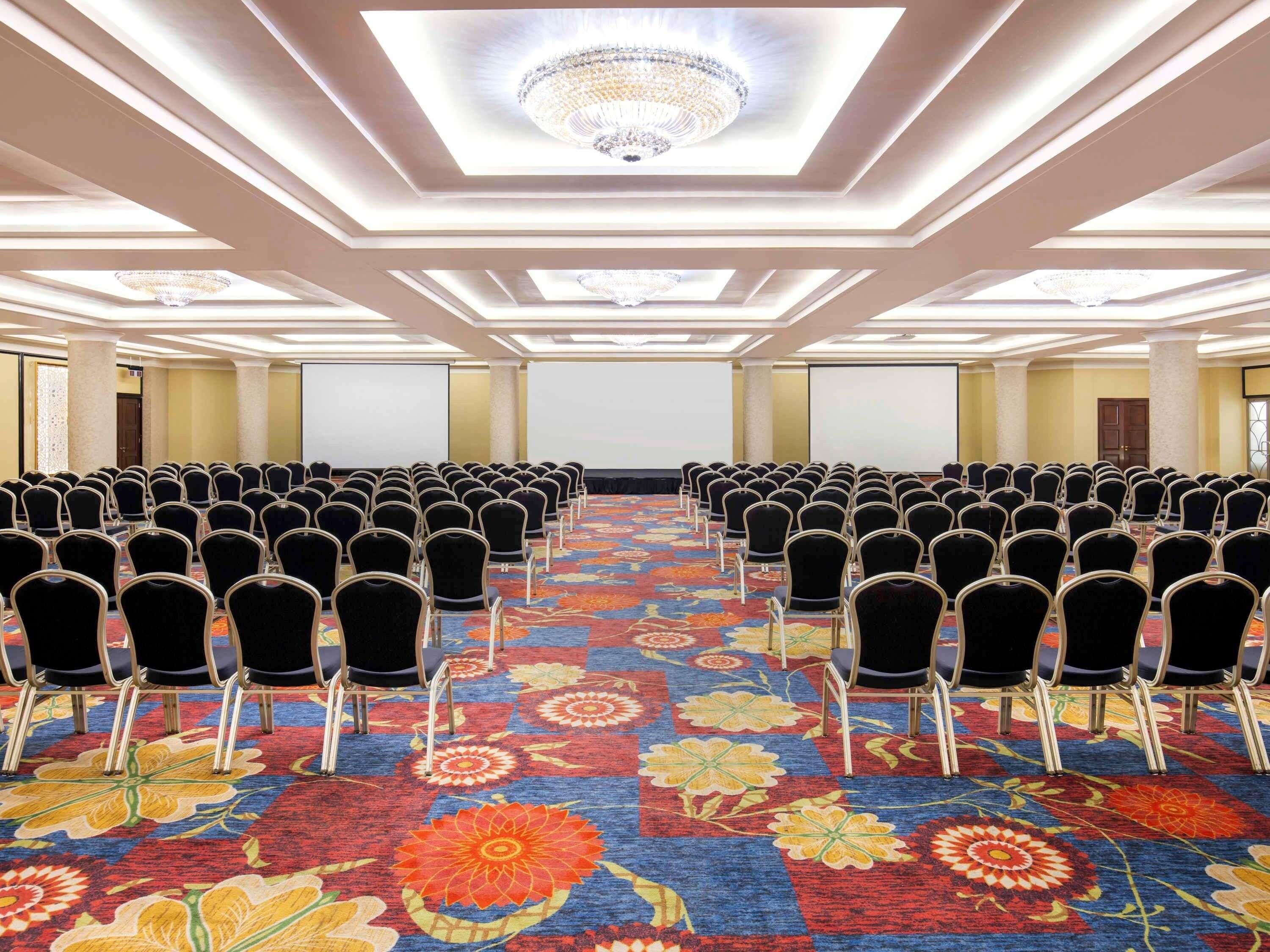 Uma sala de conferência com fileiras de cadeiras, piso coberto por carpete e um candelabro.
