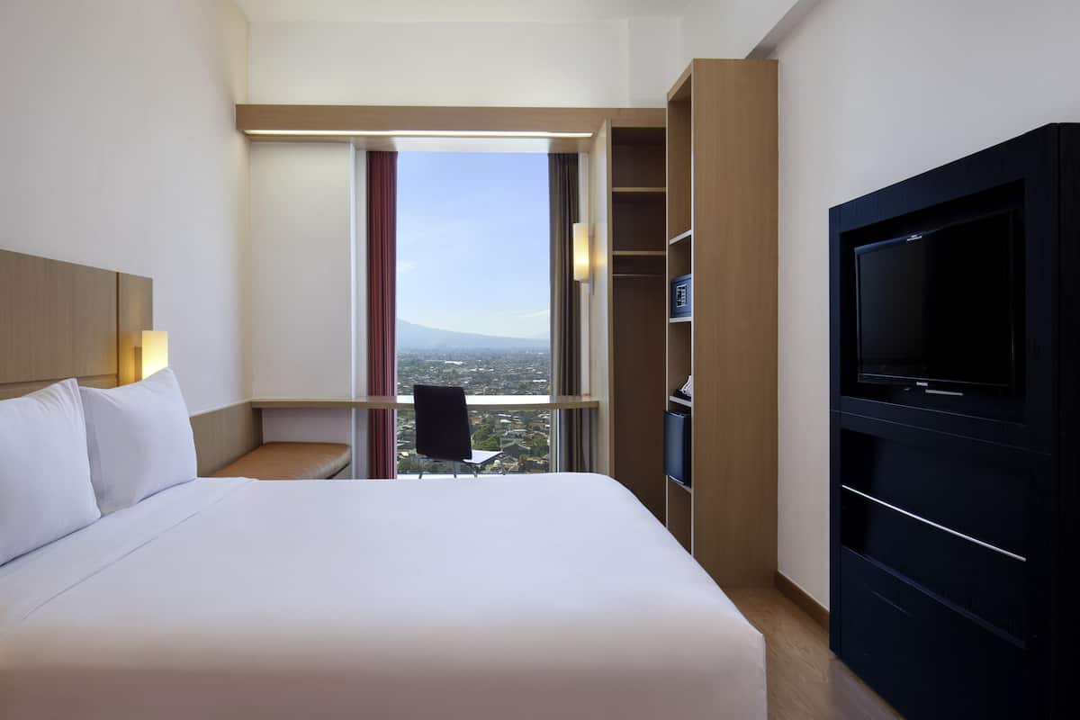 Ein Hotelzimmer mit einem großen Bett, einem Fernseher, einem Kleiderschrank und Blick auf die Stadt.