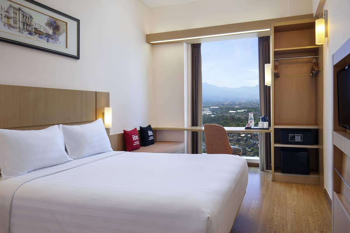 Ein Hotelzimmer mit einem großen Bett, einem Schreibtisch mit Stuhl, einem Fernseher und Blick auf eine Stadtlandschaft durch ein großes Fenster.
