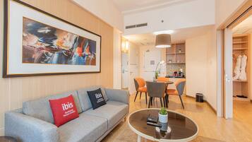 A modern hotel room with a grey sofa, a coffee table, a dining area with chairs, and a kitchen in the background.
