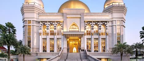 A grand building with three domes, a staircase leading to the entrance, and palm trees around.