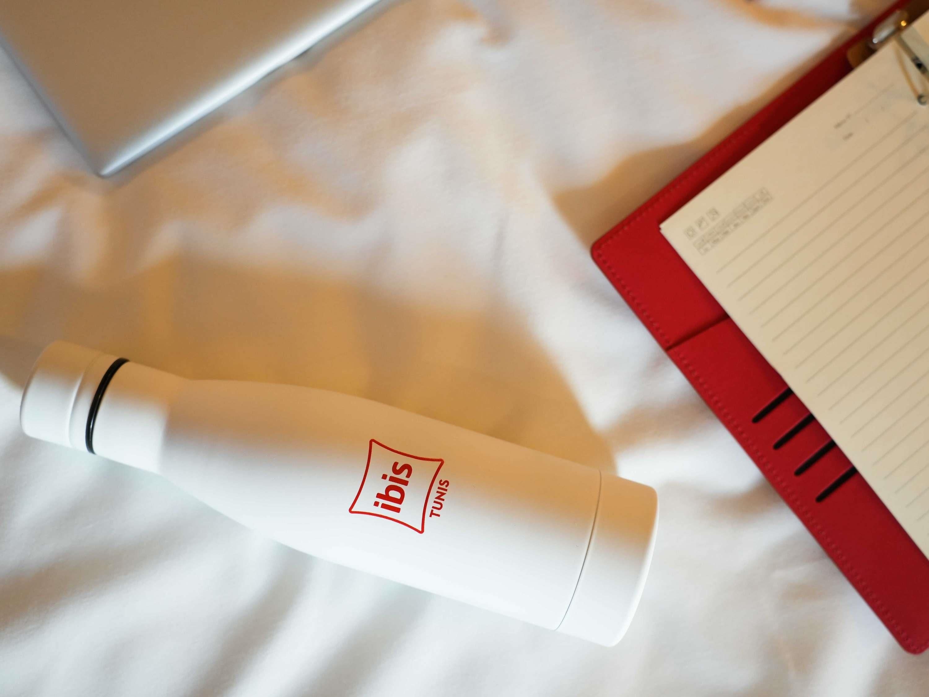 A white bottle with 'ibis' and 'Tunis' branding, a red notebook with lined pages, and a white surface.