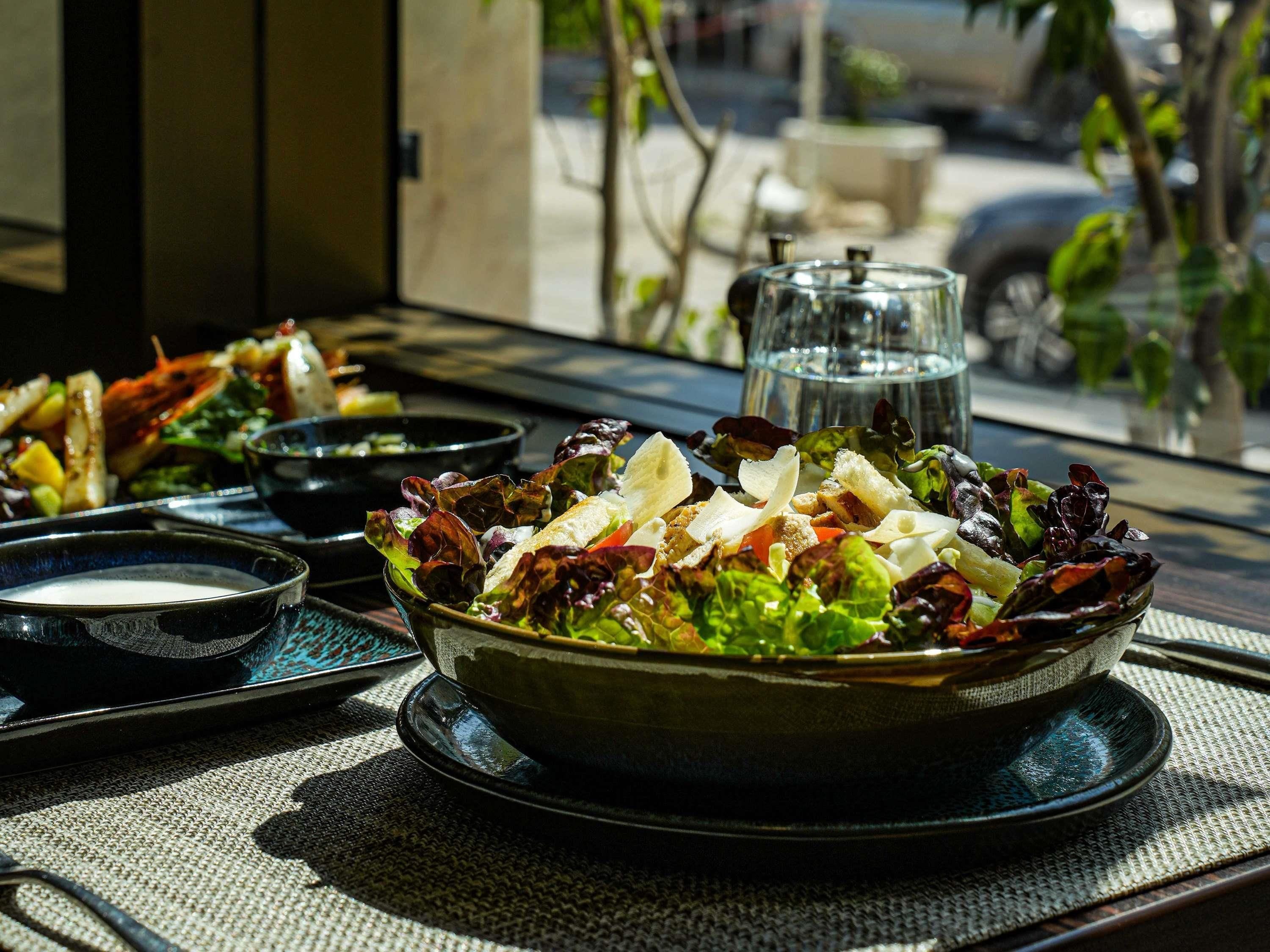 A table with a salad, bowls, and a glass container, likely containing water or a beverage, with a view of the outdoors.