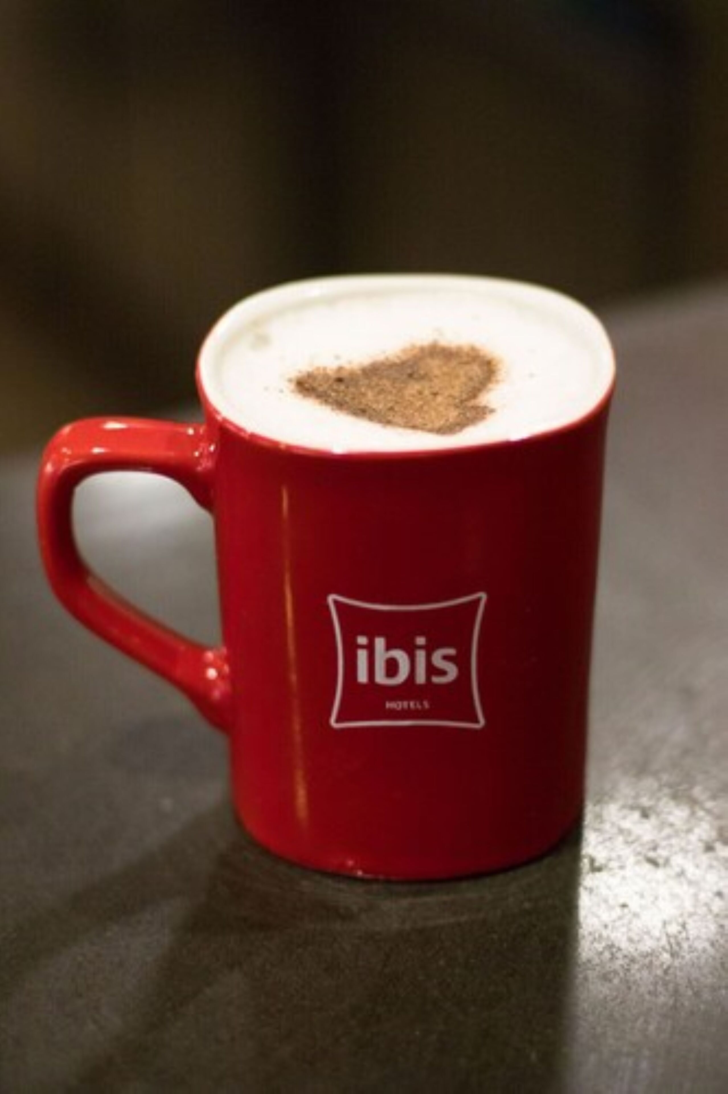 A red mug with a heart-shaped coffee foam on top and the word "ibis" on it.