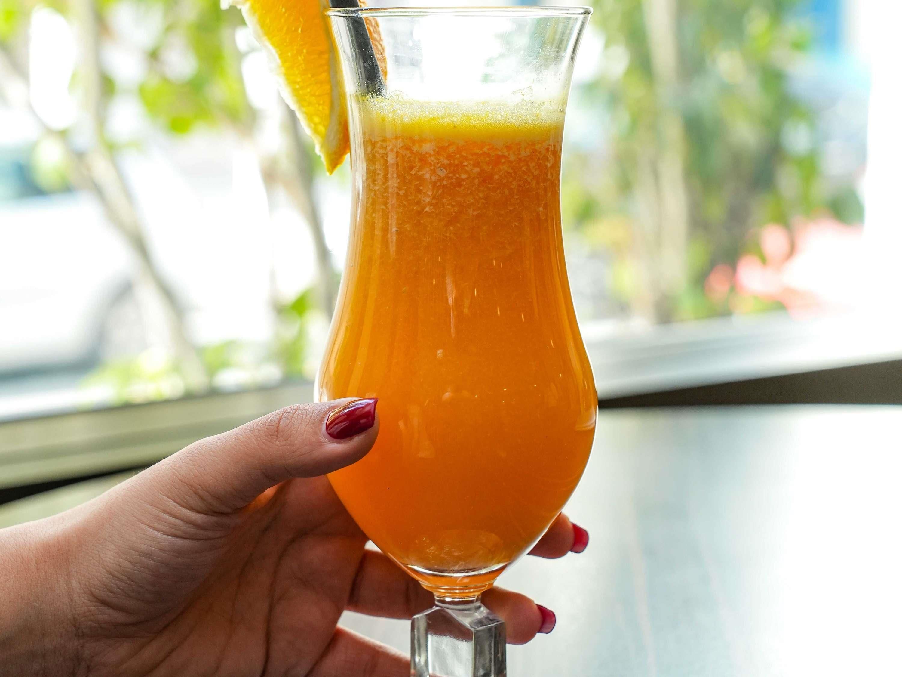 Una mano sosteniendo un vaso de jugo de naranja con una rodaja de naranja en el borde.