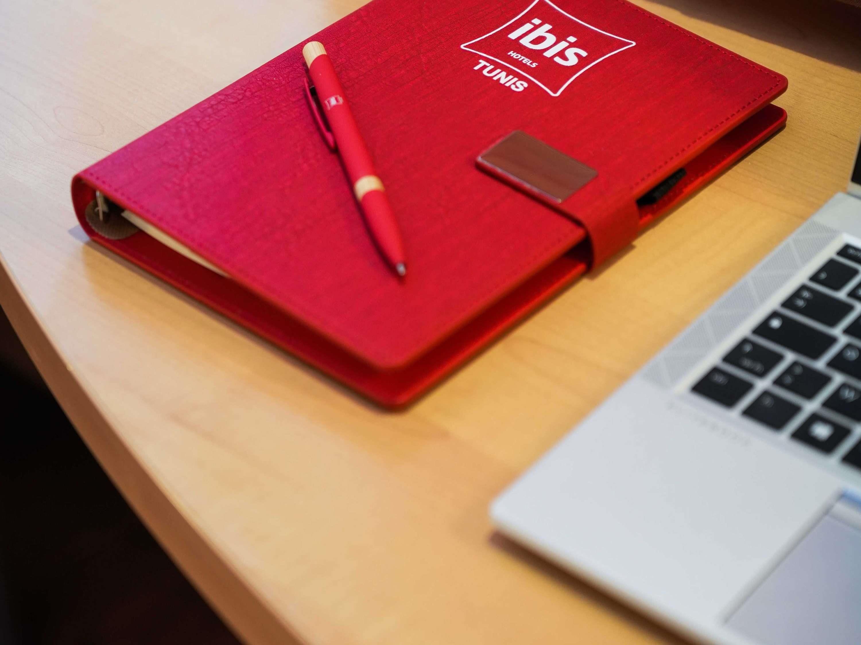 Un cuaderno rojo con la marca ibis Hotels Túnez, un bolígrafo y una computadora portátil sobre una superficie de madera.