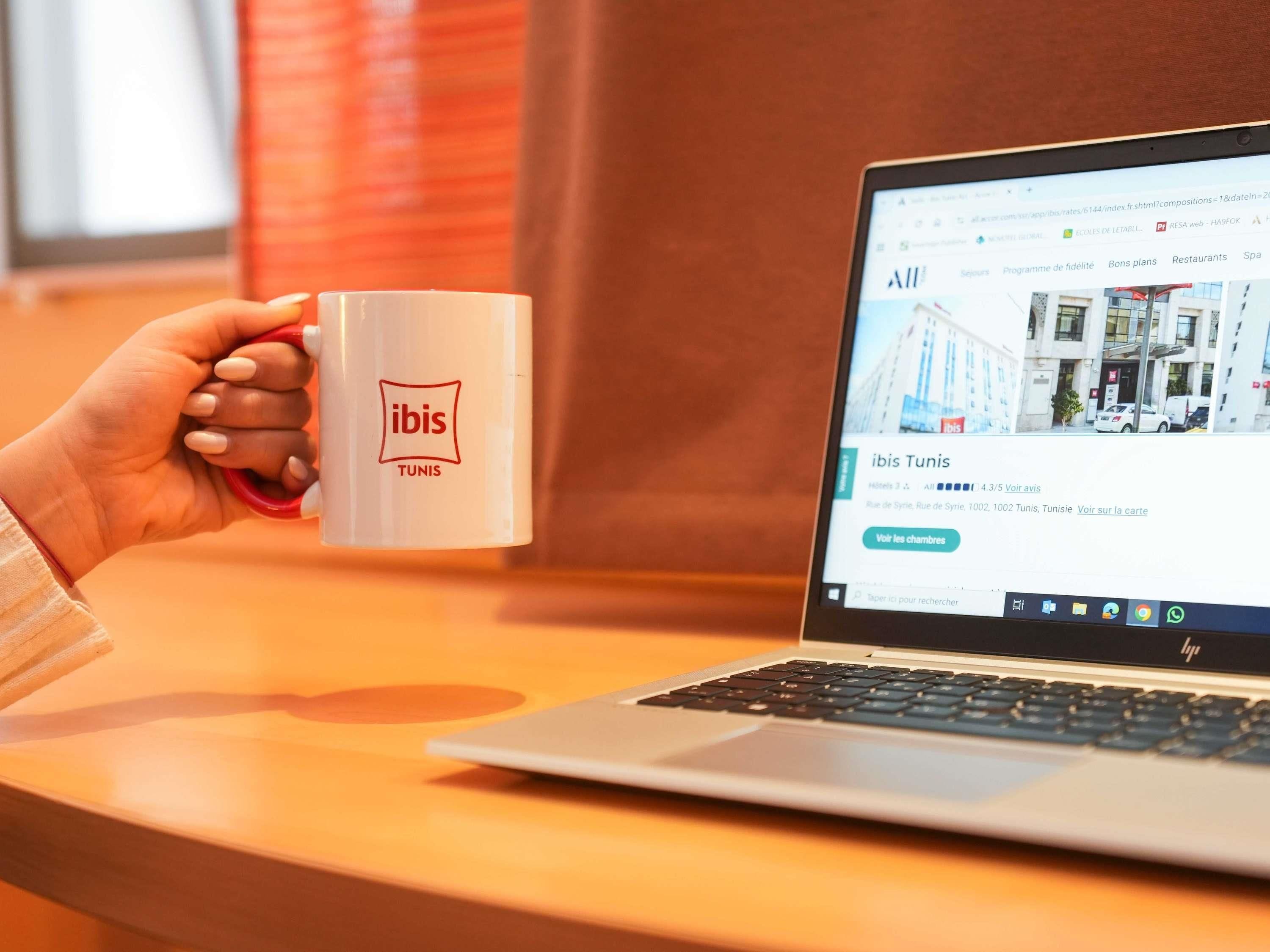 A person holding a mug with the ibis logo, sitting in front of a laptop displaying a hotel listing for ibis Tunis.