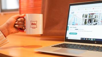 A person holding a mug with the ibis logo, sitting in front of a laptop displaying a hotel listing for ibis Tunis.