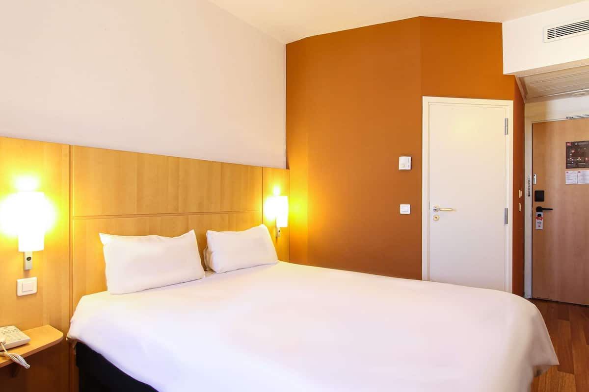Ett hotellrum med en säng, sänglampor, en telefon, en luftkonditioneringsanläggning och en dörr med lås.