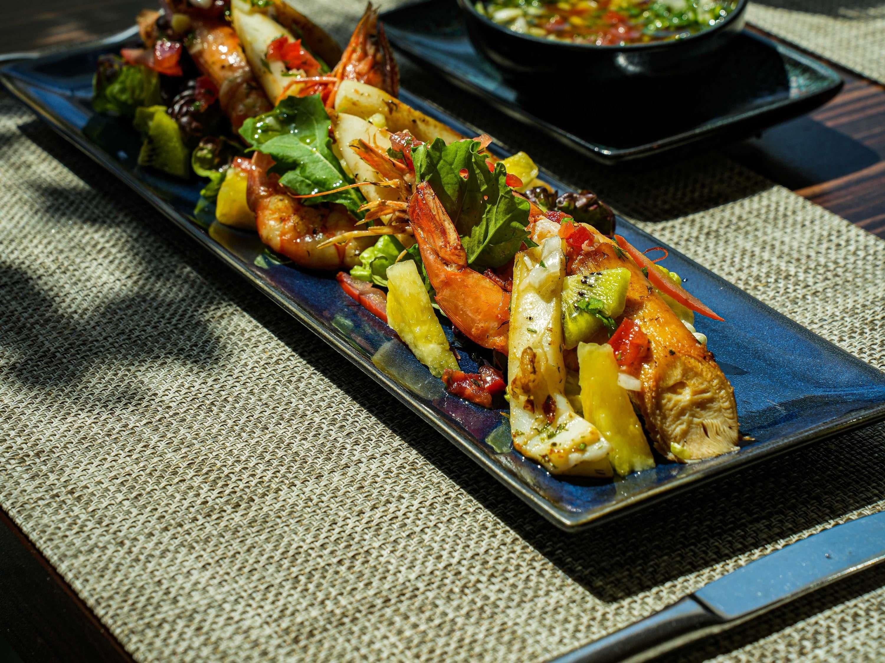 Un plato rectangular largo con camarones a la parrilla, verduras y una ensalada.