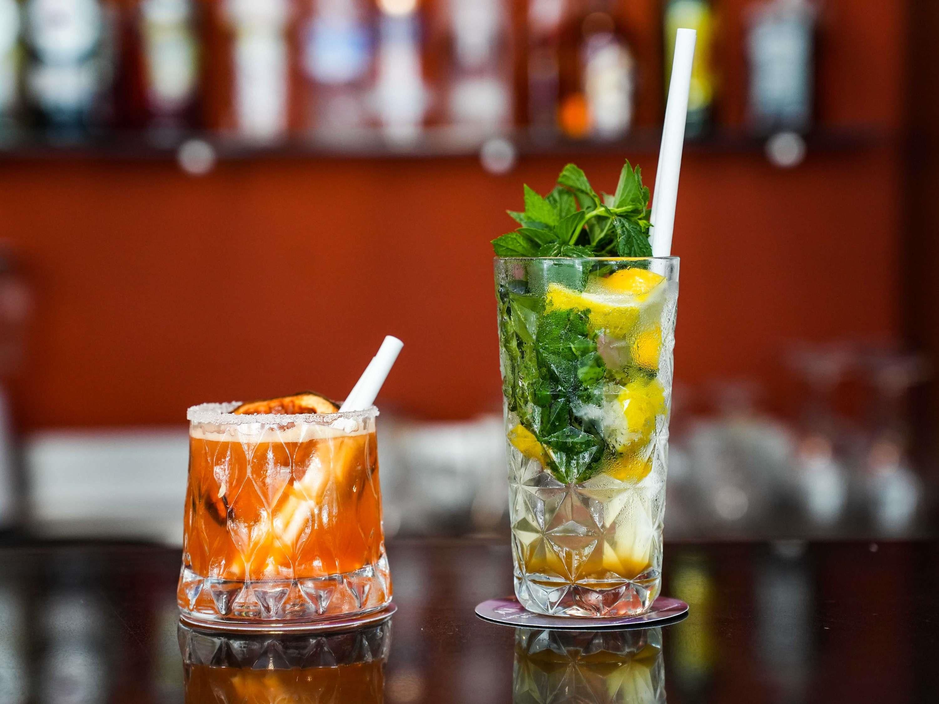 Two glasses of iced beverages on a bar counter with a blurred background of shelves.