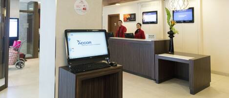 A reception area with a laptop on a stand, two receptionists, a digital screen, and a plant.