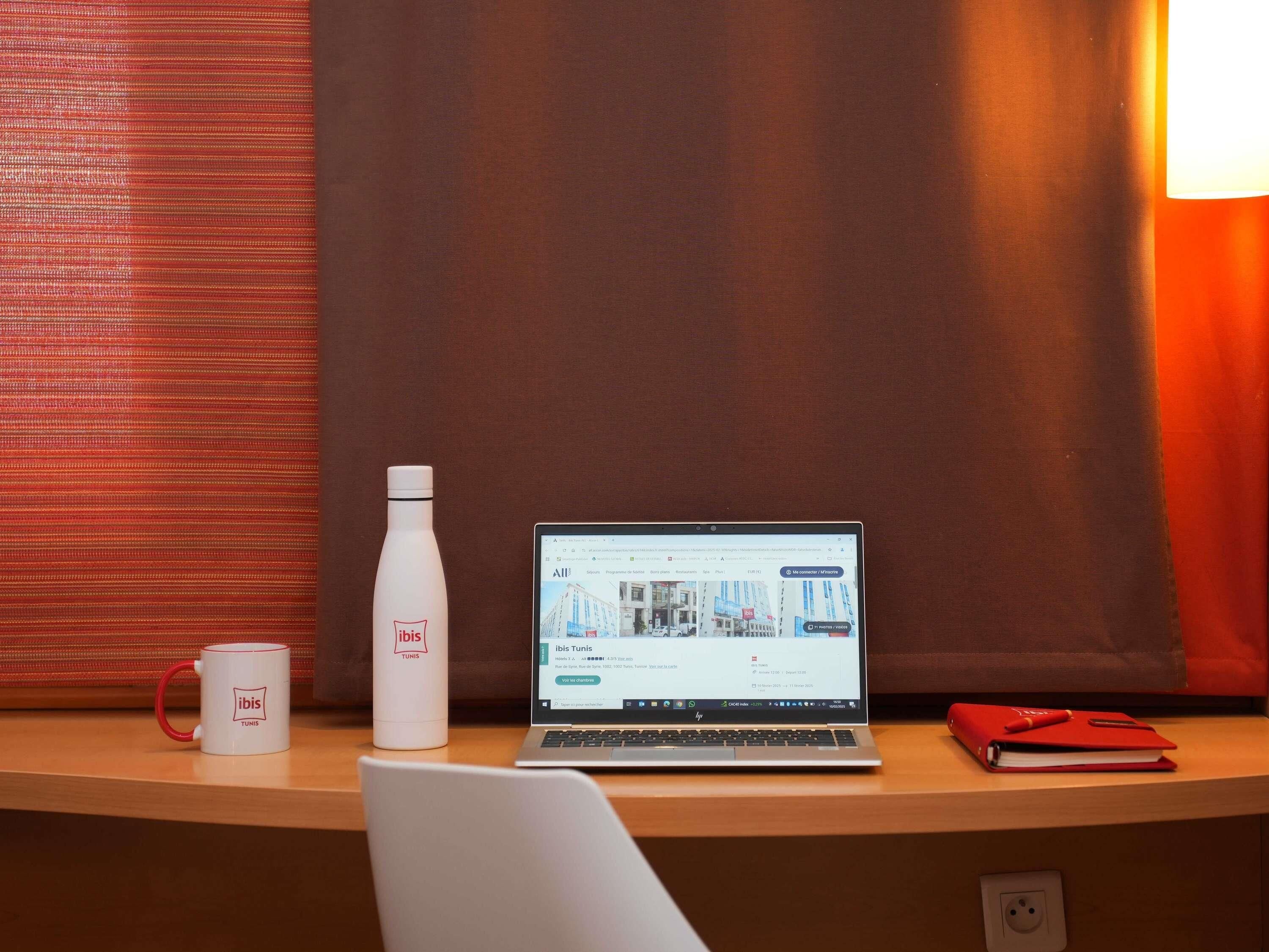 Una laptop sobre un escritorio con una taza blanca y una botella de agua, un cuaderno rojo y una silla.