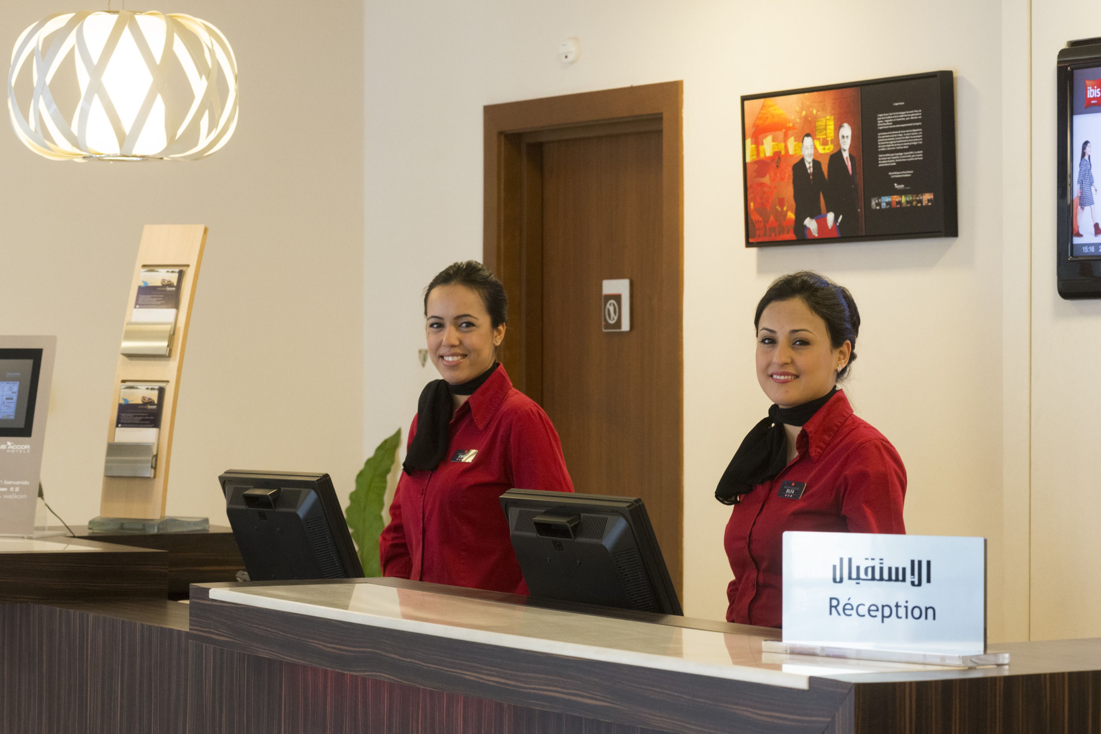 A reception desk with two receptionists in red uniforms, a sign reading "Reception" in Arabic, and a framed picture on the wall.