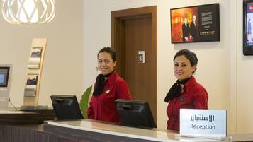 A reception desk with two receptionists in red uniforms, a sign reading "Reception" in Arabic, and a framed picture on the wall.