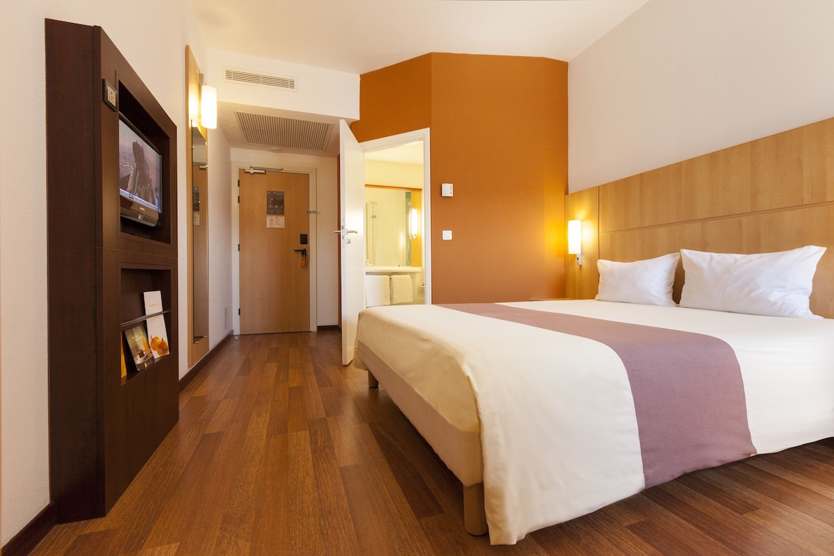Une chambre d’hôtel avec un grand lit, un plancher en bois, un téléviseur posé sur une étagère et une salle de bain visible à travers une porte ouverte.