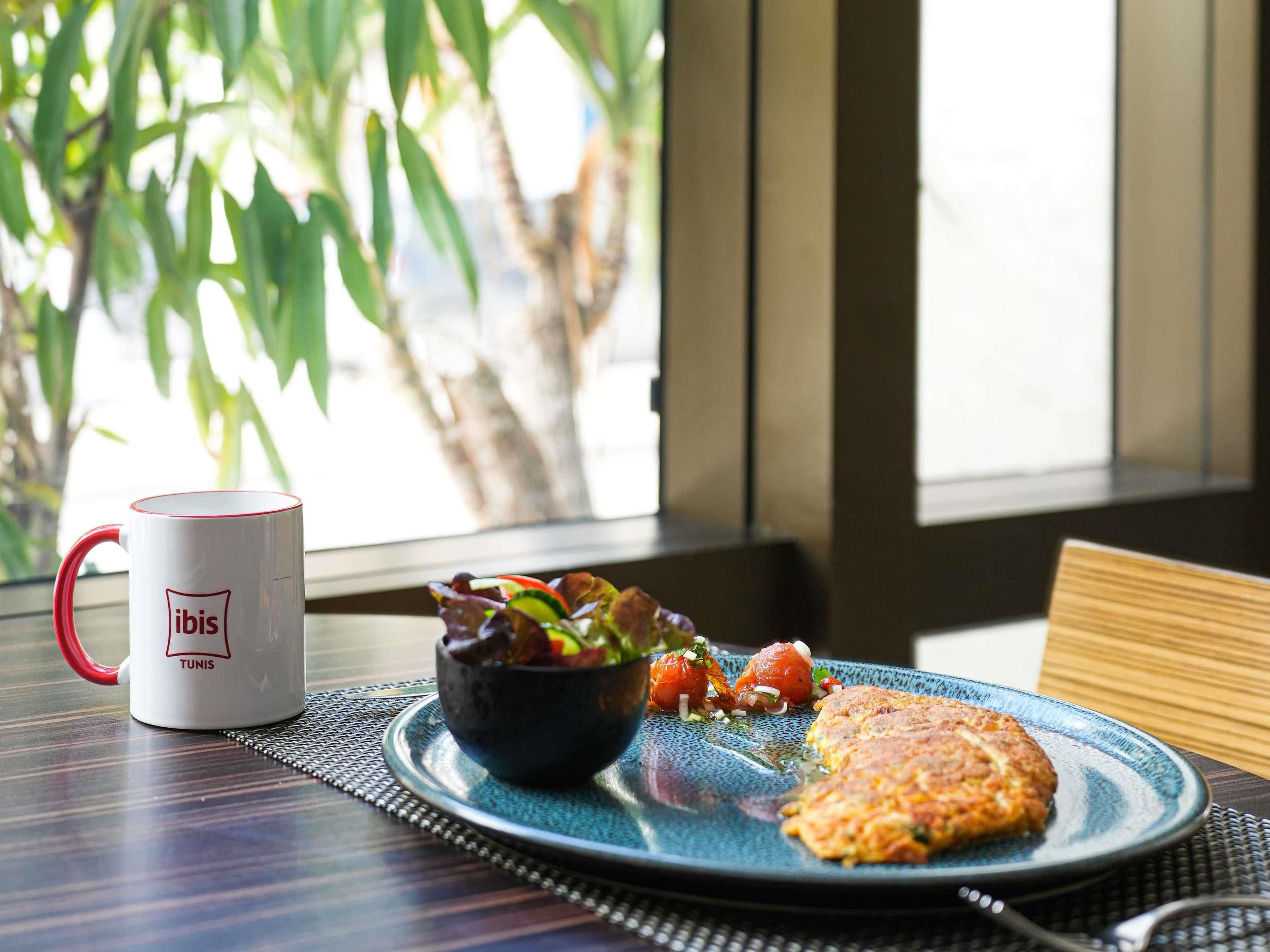 Una mesa con una taza blanca que dice "ibis TUNIS", un plato azul con ensalada y una hamburguesa, y vistas a la vegetación a través de una ventana.