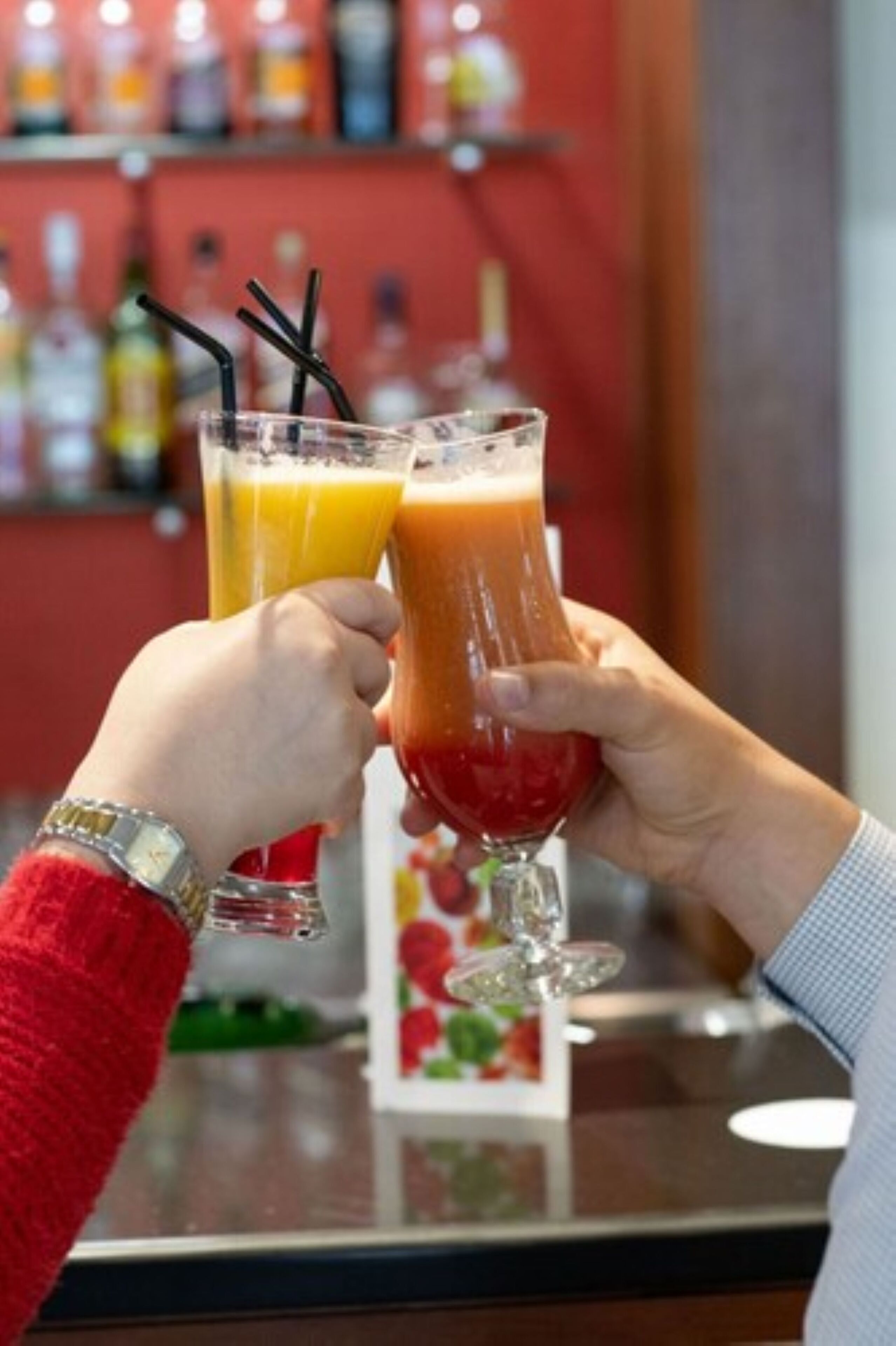 Twee personen houden glazen met een rood drankje en een geel drankje met zwarte rietjes.