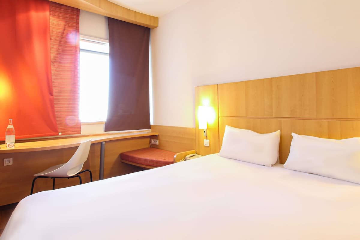 A hotel room with a bed, a desk with a chair, a telephone, and a window with curtains.