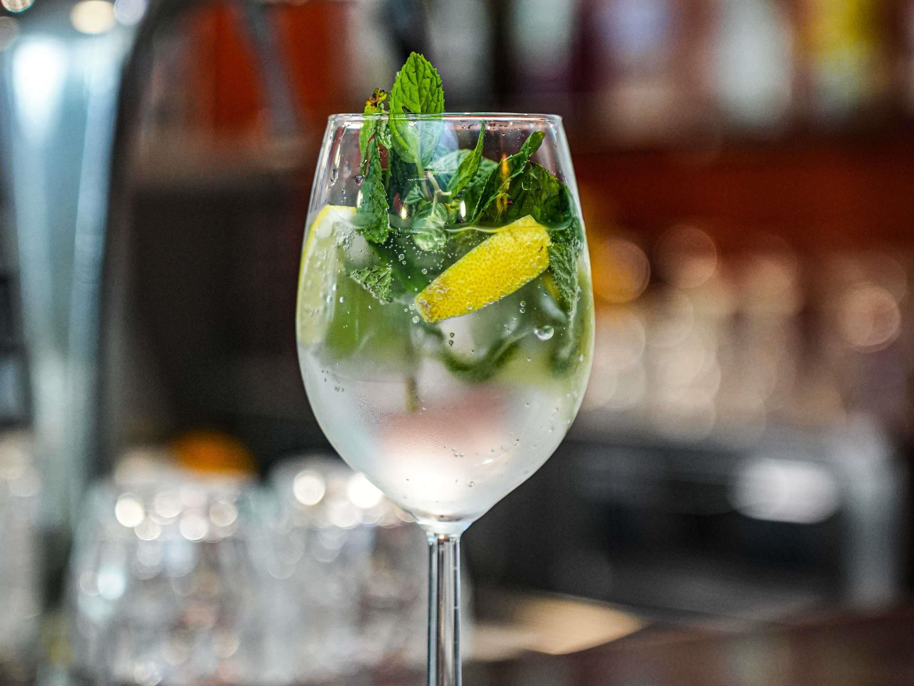 Un vaso de una bebida clara y efervescente, adornado con hojas de menta y una rodaja de limón, con un fondo borroso de bar.